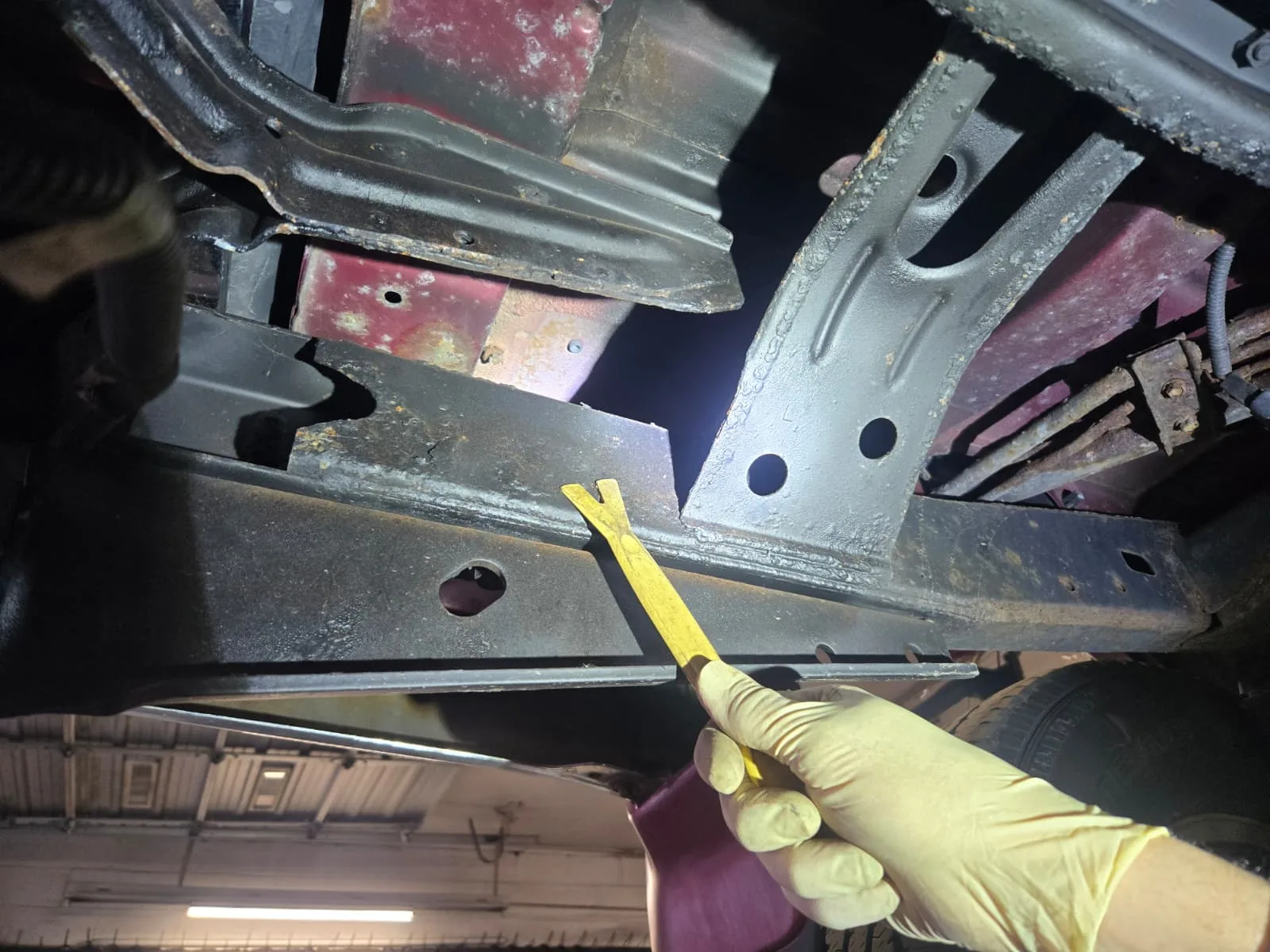 Mechanic in nitrile gloves probing a rusted vehicle frame with a pick tool during a Virginia safety inspection
